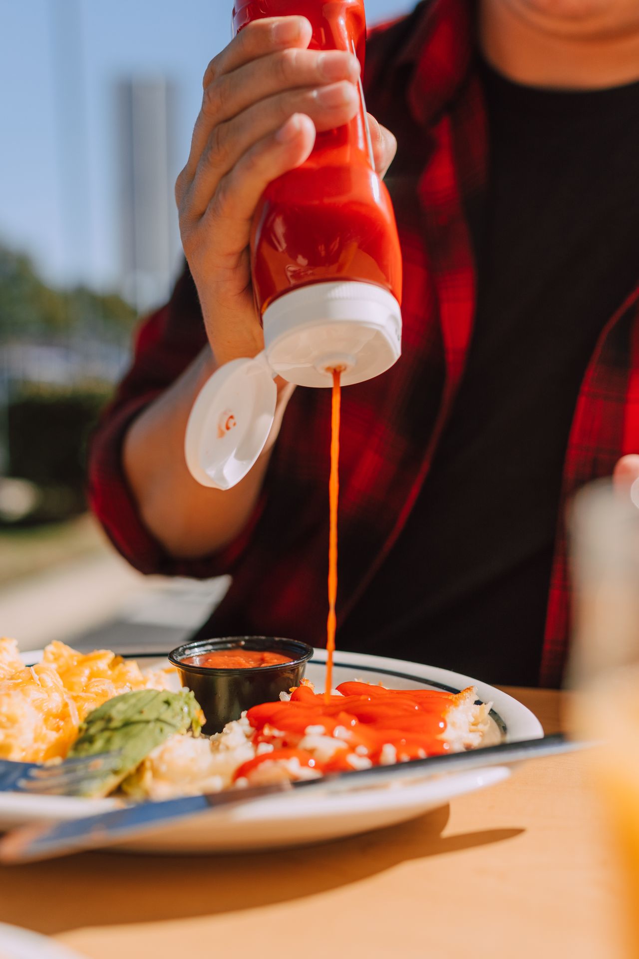 Hand pouring sriracha hot chili sauce over a rice bowl at an outdoor table